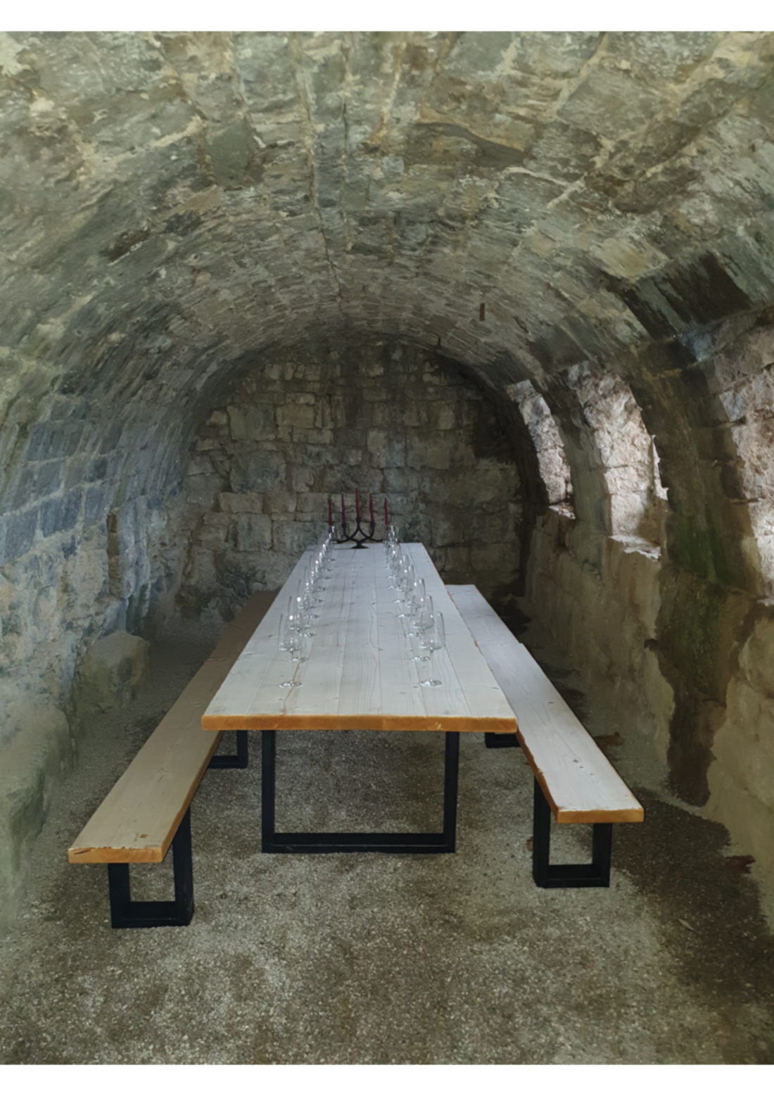 Ein langer Tisch aus Holz mit Metallbeinen steht in einem steinernen Raum mit gewölbter Decke. Auf dem Tisch sind Gläser aufgestellt. Die Wände bestehen aus grob bearbeitetem Stein und es gibt kleine Fenster, durch die Licht einfällt.