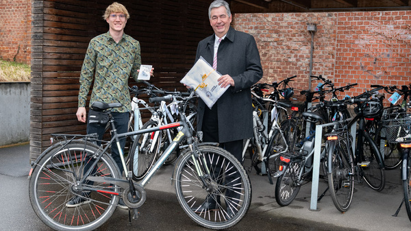 Zwei Männer stehen neben einem schwarzen Fahrrad in einer Fahrradstation. Der junge Mann hält ein kleines Gerät, während der andere Mann eine Mappe mit Unterlagen in der Hand hat. Im Hintergrund sind viele Fahrräder abgestellt.