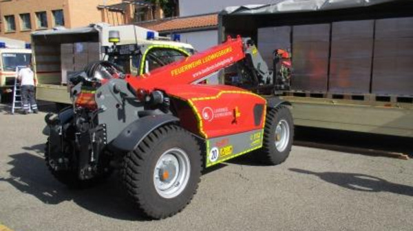 A red and black fire department telehandler is parked on a concrete area. It features large tires and a distinctive front attachment. The background includes a building and some emergency vehicles, indicating a public safety event or demonstration.