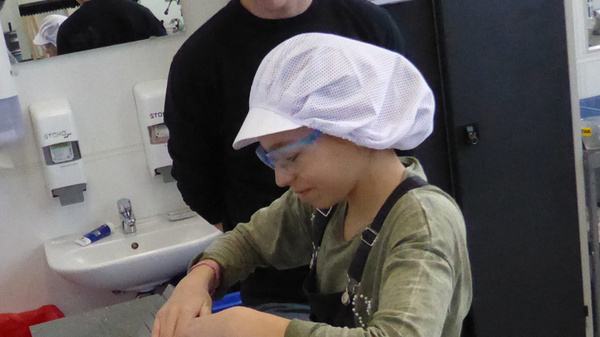 Eine Person, die eine Schutzbrille trägt, beobachtet eine zweite Person, die auf einem Tisch an einer Maschine arbeitet. Die Maschine ist blau und wird zur Bearbeitung eines Werkstücks verwendet. Beide Personen tragen geeignete Schutzkleidung.