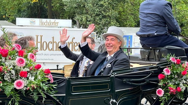 A man in formal attire and a top hat smiles and waves from a decorated horse-drawn carriage adorned with pink and white flowers. Another person sits beside him, also dressed formally. The scene suggests a festive or celebratory occasion.