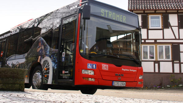 A red and black bus labeled "Stromer" is parked on a cobblestone street, with a traditional half-timbered house in the background. The bus features promotional graphics and is positioned prominently in the foreground of the image.