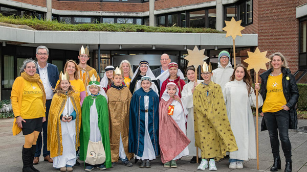 Eine Gruppe von Menschen in bunten Kostümen steht im Freien vor einem Gebäude. Einige tragen goldene Kronen und halten Sterne in den Händen. Die Atmosphäre ist fröhlich und festlich, symbolisiert durch gelbe T-Shirts mit dem Schriftzug „Sternsinger“.