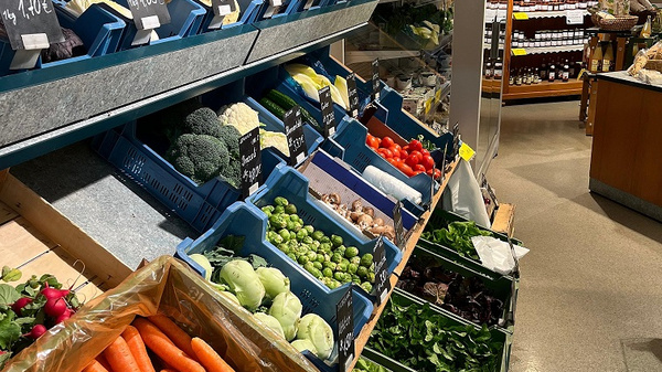 Ein Verkaufsstand in einem Lebensmittelgeschäft mit buntem Gemüse. Oben sind blaue Kisten mit Brokkoli, Blattsalat und Paprika angeordnet. Unten liegen orange Karotten und verschiedene Kohlsorten. Im Hintergrund sind Regale mit Glasbehältern gefüllt mit Konserven und anderen Lebensmitteln sichtbar.