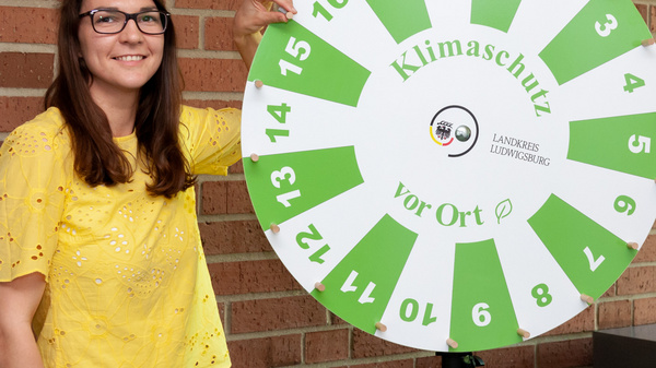 Two women stand next to a large spinning wheel labeled "Klimaschutz vor Ort," which translates to "Climate Protection On Site." The wheel features numbers and green sections. The background consists of a brick wall, emphasizing the setup for a possibly educational or promotional event.