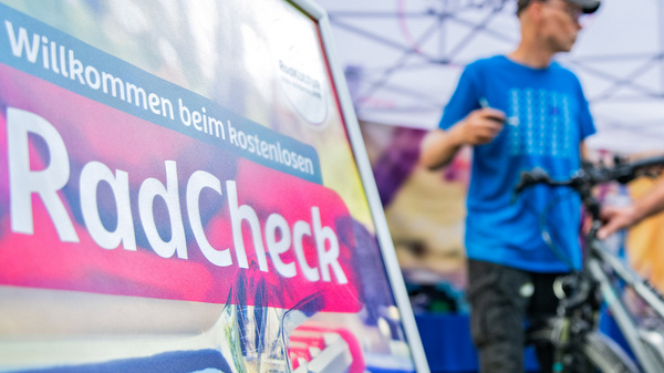 Ein Schild mit der Aufschrift „Willkommen beim kostenlosen RadCheck“ steht im Vordergrund. Im Hintergrund sind zwei Personen zu sehen, die Fahrräder überprüfen, während ein Zelt mit weiteren Informationen im Hintergrund steht.