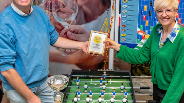 Zwei Personen stehen neben einem Tischfußballspiel. Eine Person hält einen Pokal in der Hand, während die andere eine Urkunde präsentiert. Im Hintergrund sind eine Grafik und weitere Personen beim Feiern sichtbar. Die Atmosphäre wirkt festlich und sportlich.