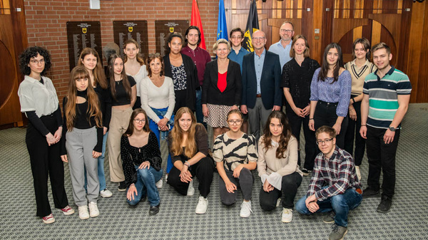 Eine Gruppe von 22 Menschen steht gemeinsam in einem Raum. Sie lächeln und posieren, einige tragen formelle Kleidung. Im Hintergrund sind Flaggen und Wappen zu sehen, die auf eine offiziellen Anlass hinweisen. Die Szene vermittelt ein Gefühl von Gemeinschaft und Beisammensein.