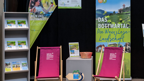 Eine Messeausstellung mit Informationsständen über das BScrolled-Region Bottenwartal. Zu sehen sind Präsentationsmaterialien, Prospekte und zwei Liegestühle. Die Farben sind einladend und harmonisch, mit Grüntönen, die die Landschaft symbolisieren. Der Hintergrund ist dunkel, um die Informationen hervorzuheben.