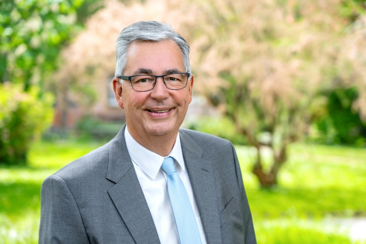 Ein lächelnder Mann mit grauen Haaren und Brille trägt einen grauen Anzug und ein hellblaues Hemd. Er steht vor einer grünen, bewachsenen Landschaft, umgeben von Bäumen und Pflanzen, und wirkt freundlich und zugänglich.