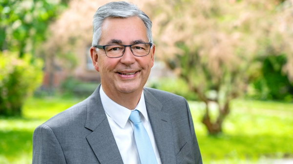 Ein lächelnder Mann mit grauen Haaren und Brille trägt einen grauen Anzug und ein hellblaues Hemd. Er steht vor einer grünen, bewachsenen Landschaft, umgeben von Bäumen und Pflanzen, und wirkt freundlich und zugänglich.