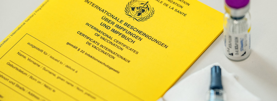 Header Bild: Ein gelbes Dokument mit der Aufschrift "Internationale Bescheinigungen über Impfungen" liegt neben einer Spritze und einer Impfstoffflasche. Es handelt sich um ein internationales Impfzertifikat, das zur Dokumentation von Impfungen dient.