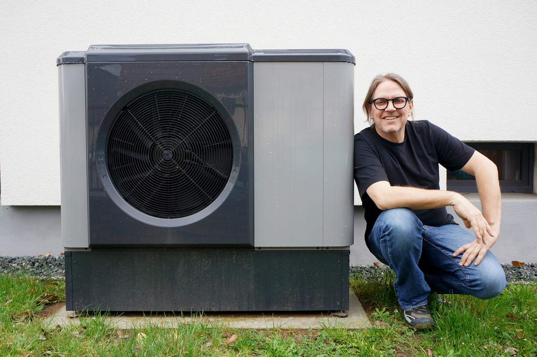 Eine Person kniet neben einer modernen Wärmepumpe, die an einer Wand installiert ist. Sie trägt eine schwarze T-Shirt und hat lange Haare. Die Umgebung ist mit Gras und kleinen Steinen gestaltet. (Text KI generiert)