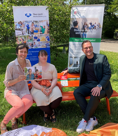 Tre persone sono sedute su una panchina all'aperto, circondate da banner informativi. Una donna mostra una foto, mentre le altre due sorridono. L'ambientazione è luminosa e verde, suggerendo un evento o una presentazione informativa.