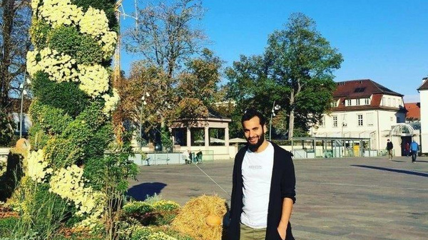 Un homme souriant se tient à côté d'une grande structure végétale décorée de fleurs blanches et vertes, entourée de plantes au sol. En arrière-plan, des bâtiments et des arbres sont visibles sous un ciel bleu clair.