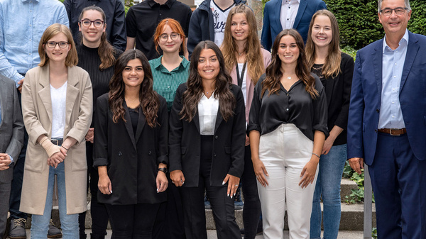 A group of 15 individuals stands on steps outside a building, arranged in three rows. They are dressed in professional attire, with a mix of suits and casual clothing. The setting features greenery in the background, creating a formal yet approachable atmosphere.