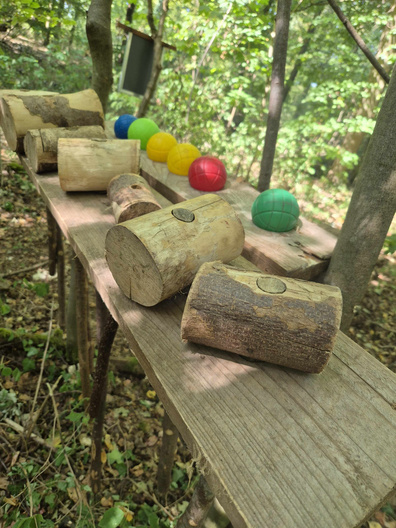 Auf einer Holztischplatte im Wald liegen mehrere Hammer aus Holz, dahinter sind bunte Bälle aufgereiht.