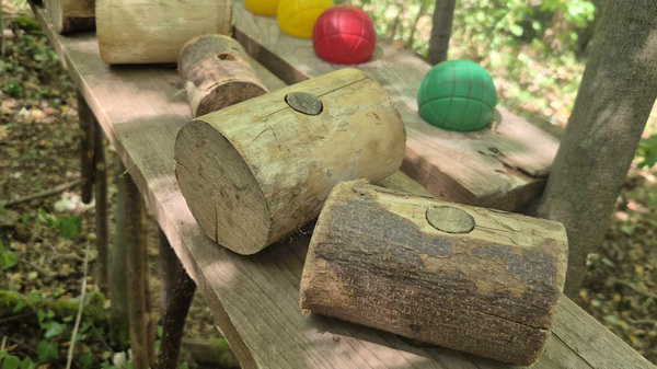 Auf einer Holztischplatte im Wald liegen mehrere Hammer aus Holz, dahinter sind bunte Bälle aufgereiht.