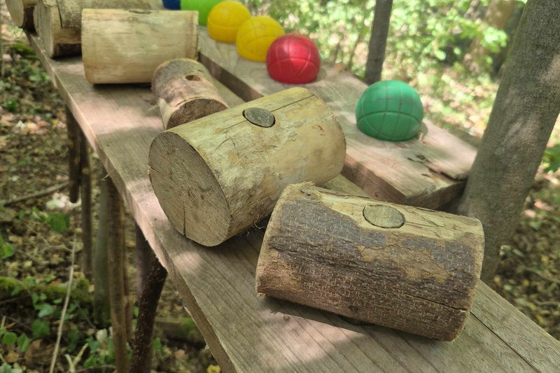 Auf einer Holztischplatte im Wald liegen mehrere Hammer aus Holz, dahinter sind bunte Bälle aufgereiht.