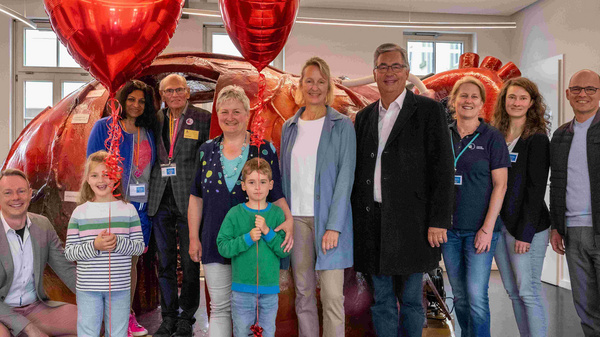 Eine Gruppe von neun Personen steht in einem Raum, umgeben von einer großen, bunten Skulptur. Einige halten Luftballons in Herzform. Die Teilnehmer sind freundlich und lächeln, was eine festliche Atmosphäre vermittelt.