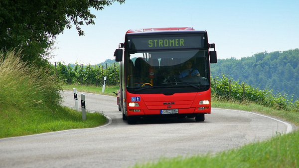 A red bus labeled "STROMER" approaches along a winding road lined with greenery. The bus is on its route, showcasing a rural landscape in the background, with trees and hills visible under a clear blue sky.