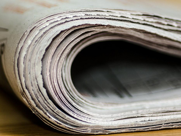 Ein zusammengefalteter Zeitungsstapel liegt auf einem Tisch. Die Seiten sind leicht gewellt und zeigen eine strukturierte Oberfläche. Die Farben der Zeitung sind dezent und neutral, und der Fokus liegt auf der Textur und Form des Papiers.