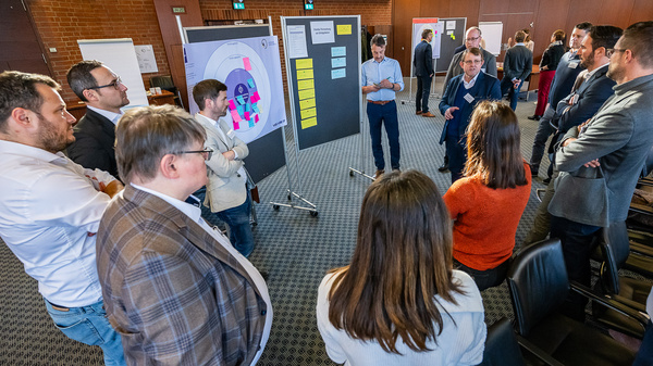 Eine Gruppe von Menschen steht in einem Raum um ein Flipchart herum, auf dem bunte Diagramme und Notizen zu sehen sind. Einige Personen diskutieren aktiv, während andere aufmerksam zuhören. Der Raum hat eine professionelle Atmosphäre, geeignet für ein Seminar oder einen Workshop.
