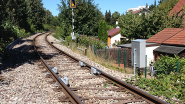 Blick auf eine kurvige Bahnstrecke, die von Schienen und Kies umgeben ist. Im Hintergrund sind Bäume und Wohnhäuser sichtbar. Ein Signal steht am Rand der Gleise. (Text KI generiert)