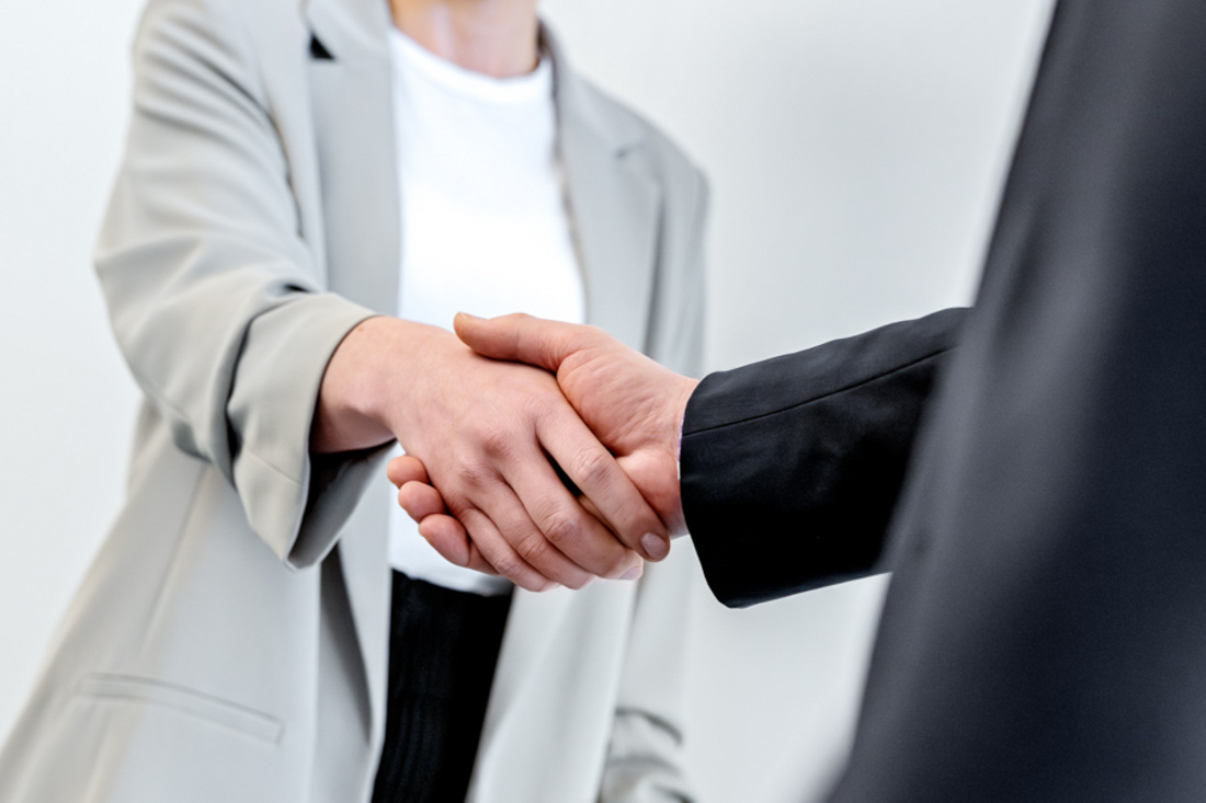 Zwei Personen schütteln sich die Hände, was ein Zeichen für eine geschäftliche Vereinbarung oder einen guten Austausch darstellt. Die Person auf der linken Seite trägt einen hellen Anzug, während die Person auf der rechten Seite einen dunklen Anzug trägt.