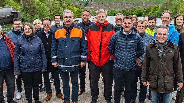 Eine Gruppe von ca. 20 Personen steht am Ufer eines Flusses, umgeben von begrünten Hügeln. Die Anwesenden tragen unterschiedliche Freizeitkleidung, lächeln und schauen in die Kamera. Die Szene vermittelt eine freundliche und gesellige Atmosphäre.