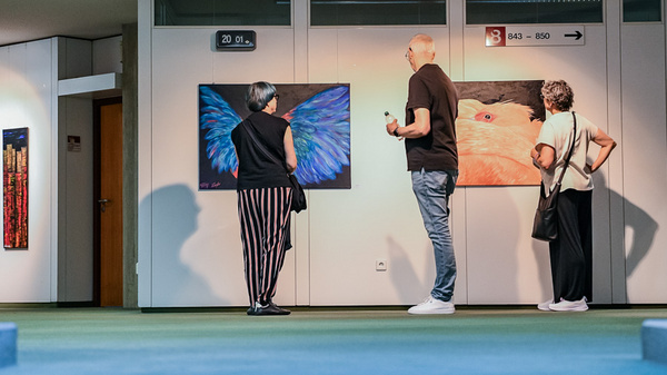 Drei Personen betrachten an einer Wand hängende Kunstwerke in einer Galerie. Eine Frau mit grauen Haaren steht links, während ein Mann in der Mitte mit einer Hand gestikuliert. Eine weitere Frau rechts schaut ebenfalls die Bilder an. Die Atmosphäre ist entspannt und kulturell.