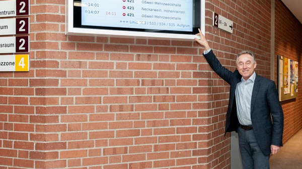 A man stands beside a digital display mounted on a brick wall, pointing towards it. The screen shows schedules for various sessions or activities, including times and locations. Nearby, directional signs indicate rooms and areas, with a specific focus on directions to a conference zone.