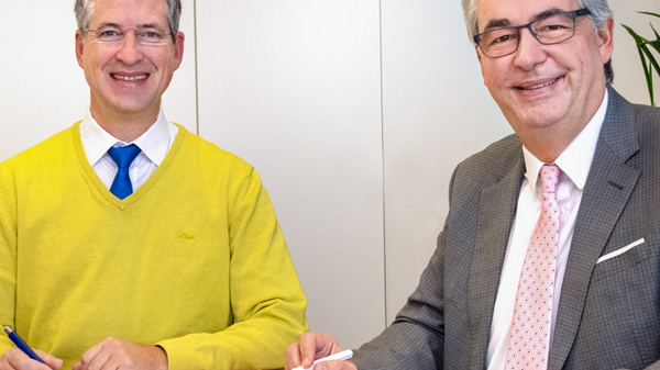 Two men sit at a wooden table, smiling as they sign documents. One man wears a bright yellow sweater and holds a pen, while the other is dressed in a gray suit with a pink tie. A red plant is visible on the table.