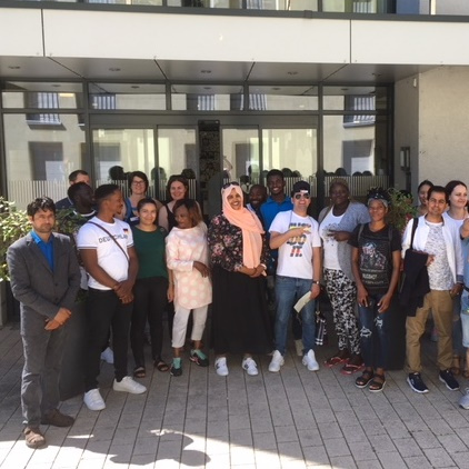 Eine Gruppe von etwa 20 Menschen steht lächelnd vor einem Gebäude. Sie sind vielfältig gekleidet und zeigen ein Gefühl von Gemeinschaft. Im Vordergrund sind auch einige Kinder zu sehen, die in den Armen ihrer Eltern gehalten werden.