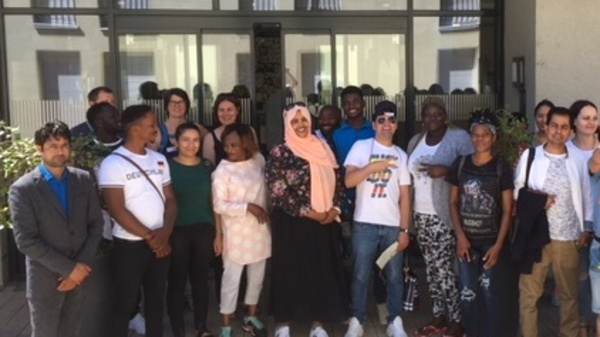 A diverse group of individuals stands together outside a building, smiling and posing for the camera. The group includes men, women, and children, wearing casual clothing and displaying a sense of community and togetherness.