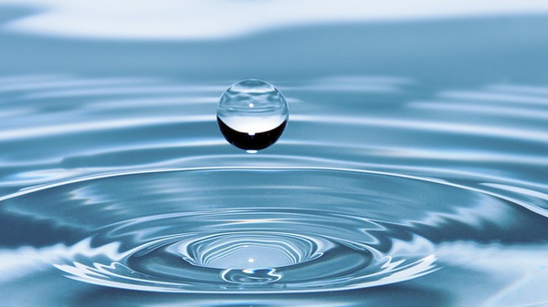 A single water droplet hovers above the surface of a calm body of water. The droplet is perfectly round, creating ripples as it prepares to land, with soft waves radiating outward, highlighting the tranquility of the scene.