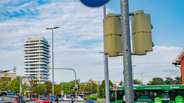 Das Bild zeigt eine Verkehrsinszenierung an einem Kreisverkehr mit Verkehrsschildern für Radfahrer und Fußgänger. Im Hintergrund sind Busse, Autos und ein modernes Gebäude zu sehen, unter blauem Himmel mit teilweise Wolken.