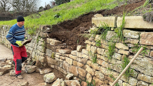 Ein Mann in gestreifter Kleidung arbeitet an einer Steinmauer. Er hält einen Schaufel und entfernt Erdreich und Steine, während er die Mauer repariert. Im Hintergrund sind Bäume und ein Hang mit frischem Gras zu sehen.