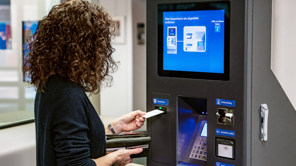Eine Person steht vor einem Automaten, während sie eine Karte in der Hand hält. Auf dem Bildschirm des Automaten sind Anweisungen zur Nutzung abgebildet. Die Umgebung wirkt modern und gut beleuchtet.
