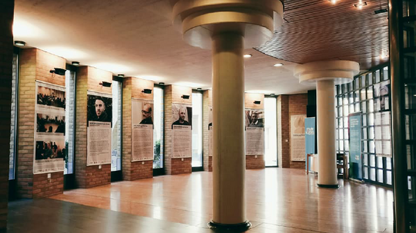 An interior space features large columns and wooden ceiling panels. Surrounding the room are several exhibition panels with text and images. Natural light filters in through large windows, illuminating the area and creating a warm, inviting atmosphere for visitors.