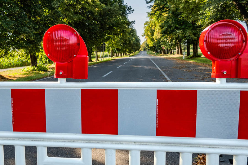 Eine Straßenabsperrung mit roten und weißen Markierungen. Im Hintergrund ist eine gerade Straße zu sehen, umgeben von Bäumen. Die Absperrung deutet darauf hin, dass der Zugang zur Straße eingeschränkt oder nicht erlaubt ist.