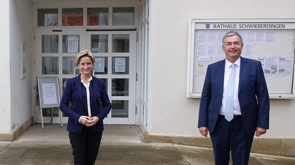 Three individuals stand outside a building labeled "Rathaus." Two men are on the left and right, wearing suits, while a woman in a suit stands in the center. They appear to be posing for a photograph, with a doorway and signs visible behind them.