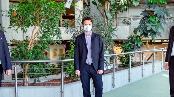 Three men, wearing face masks and professional attire, stand together in a green indoor space filled with potted plants. They are positioned in a semi-formal setting, with a modern architectural background and railing separating them from a walkway.