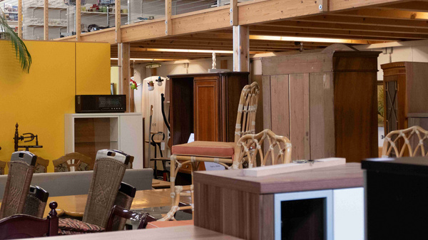 Das Bild zeigt eine Möbelbörse mit einer Vielzahl an Holzmöbeln, darunter Stühle, Tische und Schränke. Im Hintergrund sind Regale mit diversen Artikeln zu sehen, während die Wände teilweise gelb gestrichen sind. Die Einrichtung vermittelt eine gemütliche und einladende Atmosphäre.