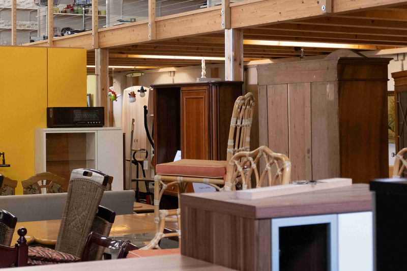 Das Bild zeigt eine Möbelbörse mit einer Vielzahl an Holzmöbeln, darunter Stühle, Tische und Schränke. Im Hintergrund sind Regale mit diversen Artikeln zu sehen, während die Wände teilweise gelb gestrichen sind. Die Einrichtung vermittelt eine gemütliche und einladende Atmosphäre.