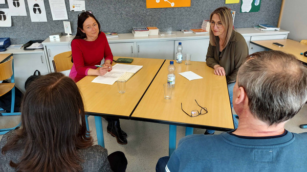Vier Personen sitzen an einem Tisch in einem Besprechungsraum. Zwei Frauen, eine in rotem Oberteil und die andere in grauem, diskutieren, während zwei andere Personen ihnen zuhören. Auf dem Tisch liegen Notizblätter und Wasserflaschen. Im Hintergrund sind organisatorische Materialien sichtbar.