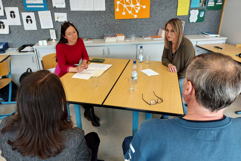 Vier Personen sitzen an einem Tisch in einem Besprechungsraum. Zwei Frauen, eine in rotem Oberteil und die andere in grauem, diskutieren, während zwei andere Personen ihnen zuhören. Auf dem Tisch liegen Notizblätter und Wasserflaschen. Im Hintergrund sind organisatorische Materialien sichtbar.