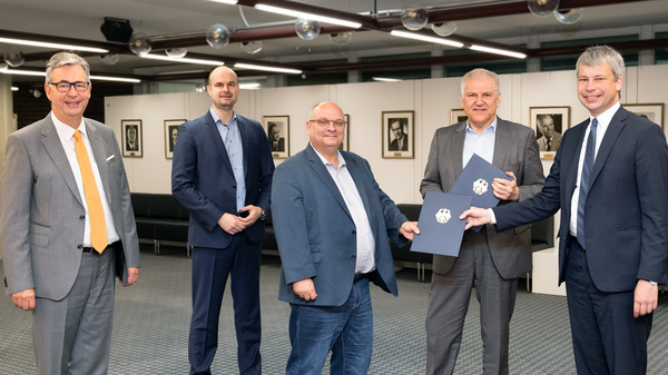 Cinq hommes en costume se tiennent debout dans une salle. L'un d'eux remet des documents à un autre. L'ambiance est formelle, indiquant une signature ou un accord. En arrière-plan, des portraits sont accrochés aux murs.