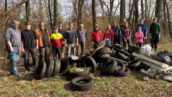 Eine Gruppe von 16 Personen steht in einem Waldgebiet neben einem Haufen Müll, der vorwiegend aus alten Reifen besteht. Die Teilnehmer tragen Arbeitskleidung und wirken stolz. Das Bild zeigt den gemeinsamen Einsatz für eine sauberere Umwelt.