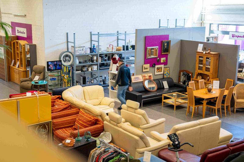 Ein Auktionshaus oder Möbelgeschäft mit verschiedenen Möbeln und Dekorationsgegenständen. Zu sehen sind Sofas, ein Esstisch mit Stühlen und Regale im Hintergrund. Menschen bewegen sich zwischen den Möbeln. (Text KI generiert)